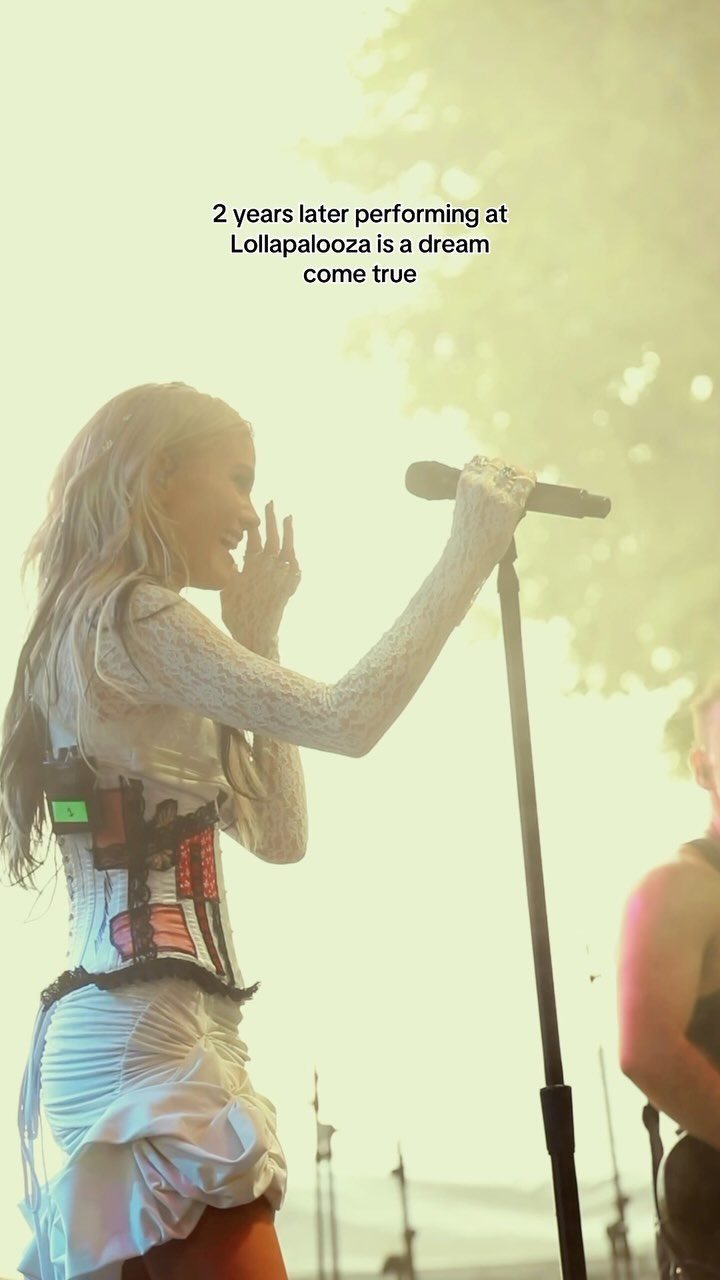 a woman is singing on stage at lollapalooza. she is wearing a corset and a white dress. she has a microphone in her hand and a microphone stand is visible in the image. there is a band member to the right of her. the text on the image says "2 years later performing at lollapalooza is a dream come true."
