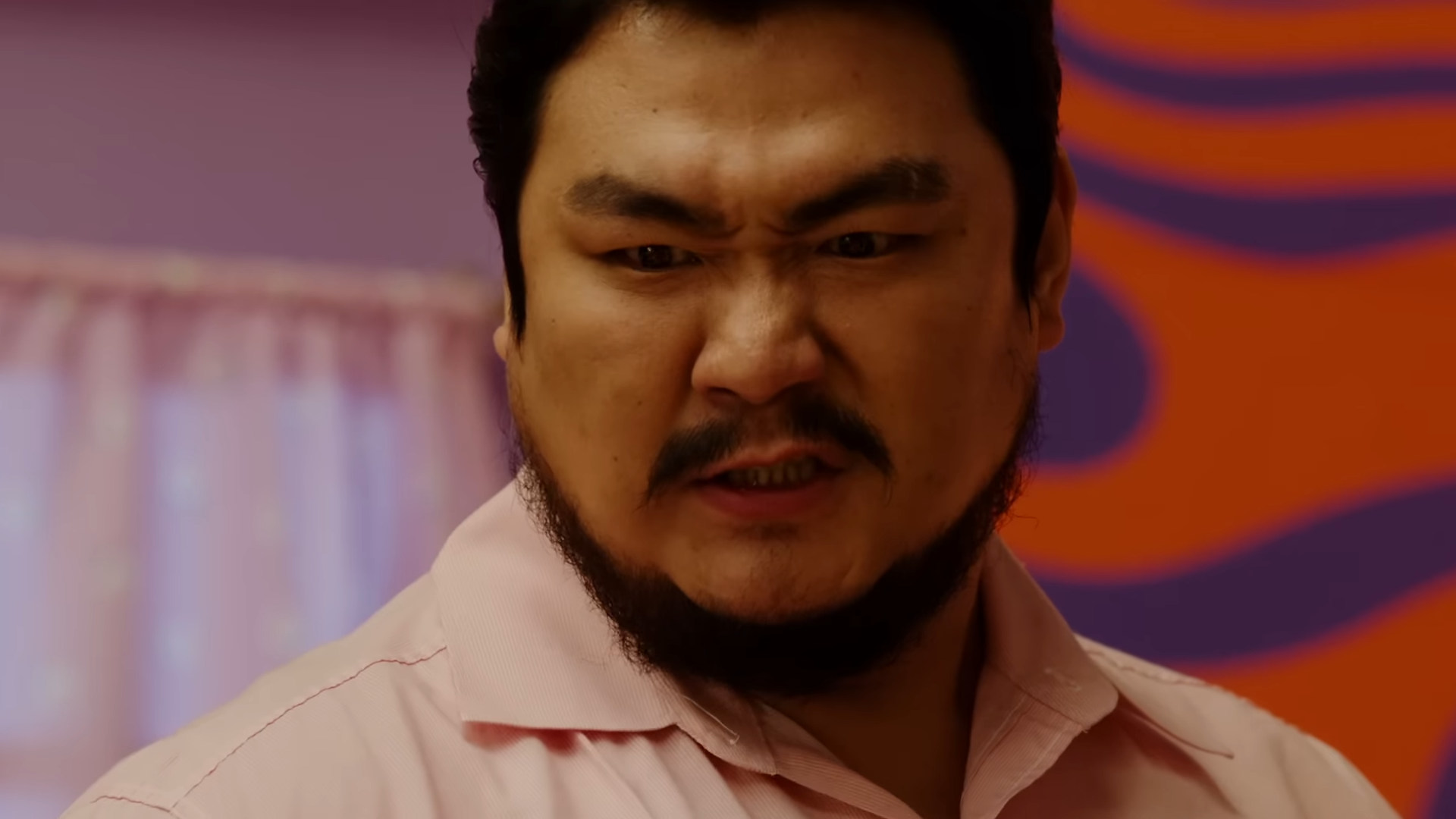 a closeup photo of a man with an angry or intense expression on his face. he has a goatee and is wearing a pink shirt. the background is out of focus and features a pink and orange pattern.