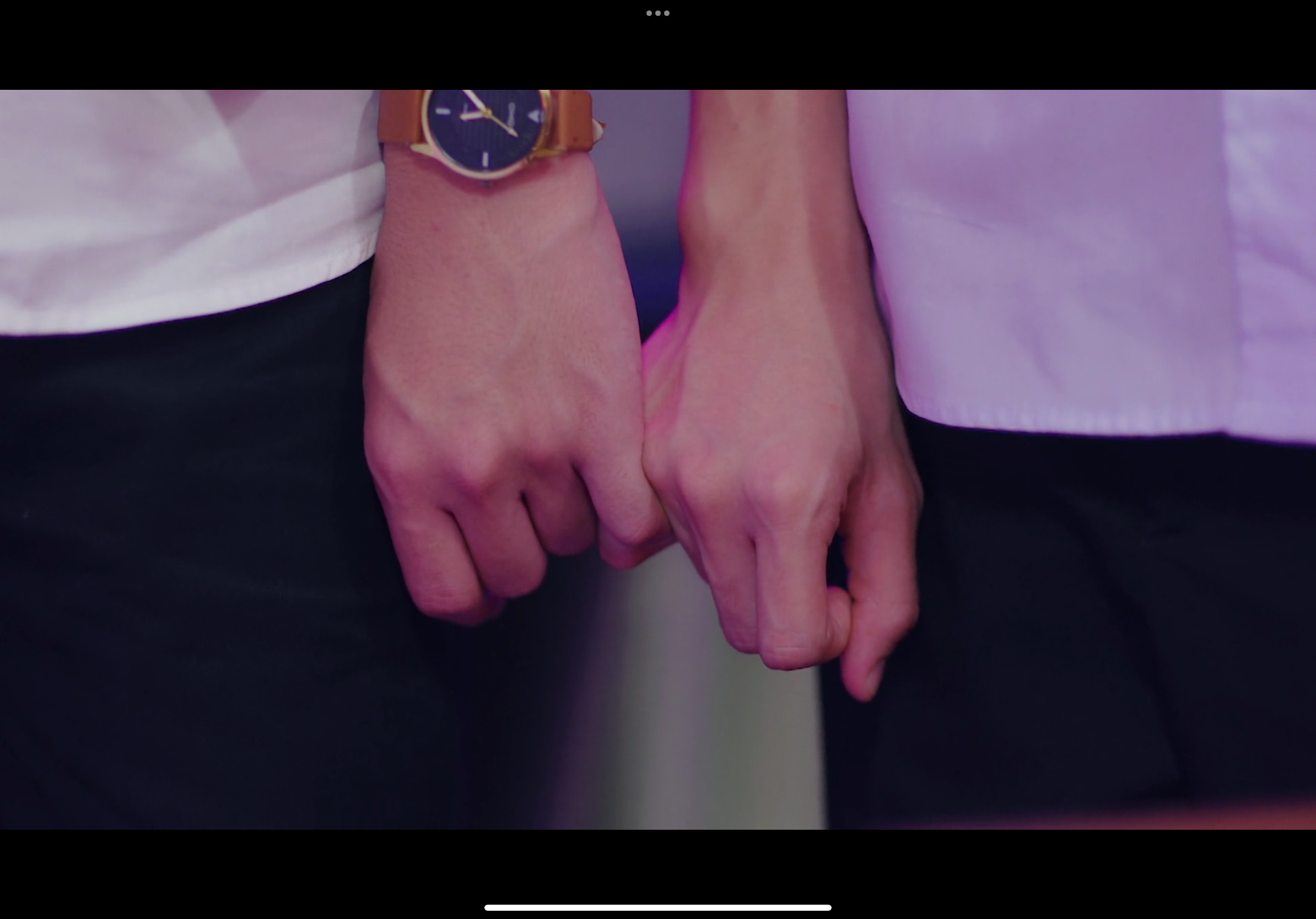 the image shows a closeup of two people holding hands with interlocked fingers. the person on the left wears a white shirt and a brown watch, while the person on the right wears a light purple shirt. the background is blurred and illuminated with pink and purple neon lighting. the focus on the hands and the vibrant colors evoke a sense of intimacy and youthful affection.
