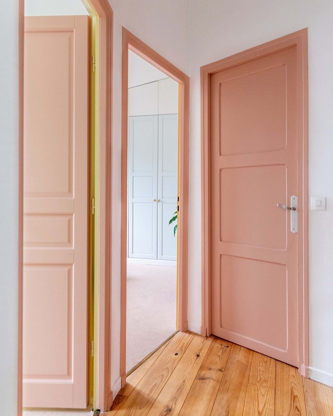the image shows a hallway with three doorways. the doors and doorframes are painted in a salmon pink color. the floor is made of wooden planks. a wardrobe can be seen through one of the doorways.