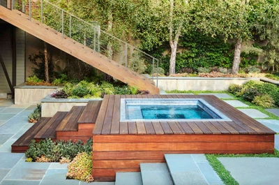 the image shows a modern outdoor hot tub built into a wooden deck with builtin steps leading down to a patio. the hot tub is surrounded by lush landscaping that includes various plants, succulents, trees, and shrubs. the overall style is minimalist and contemporary, creating a serene and relaxing atmosphere. the design integrates seamlessly with the surrounding architecture and garden, enhancing the natural beauty of the setting. the stone patio provides a clean contrast to the rich tones of the wood.