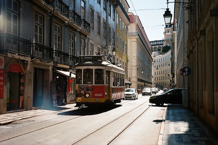 the image depicts a sunny lisbon street scene, featuring a vintage red and yellow tram (streetcar) traveling along tram tracks. the tram is labeled "m. moniz".  the street is narrow, lined with traditional buildings with characteristic portuguese azulejo tiled facades and balconies.  cars are parked along the sides of the road. the architecture appears old and historic, creating a picturesque, charming atmosphere.  the perspective is from a lower vantage point, looking up the street towards a hill with more buildings visible in the distance. a cocacola advertisement is visible on the side of the tram. the overall feeling is one of a classic, vibrant european city scene.
