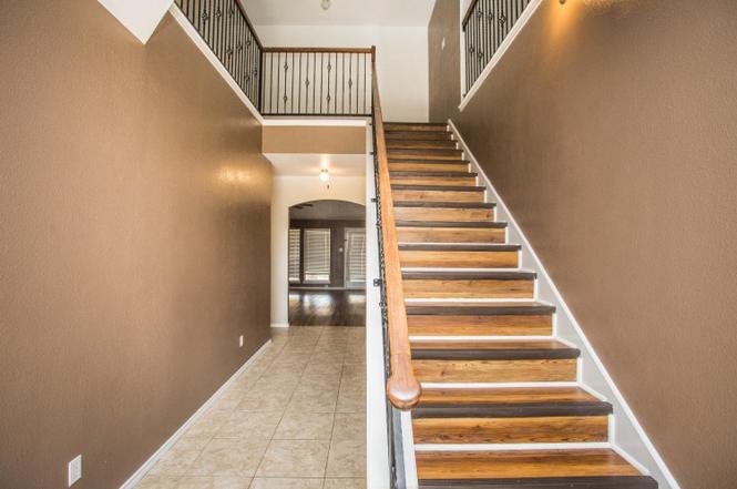 the image shows the interior of a twostory house. there is a staircase with a banister and railing going up to the second floor. the hallway has a tile floor and leads to another room. the walls are painted brown. the decor is simple and modern.
