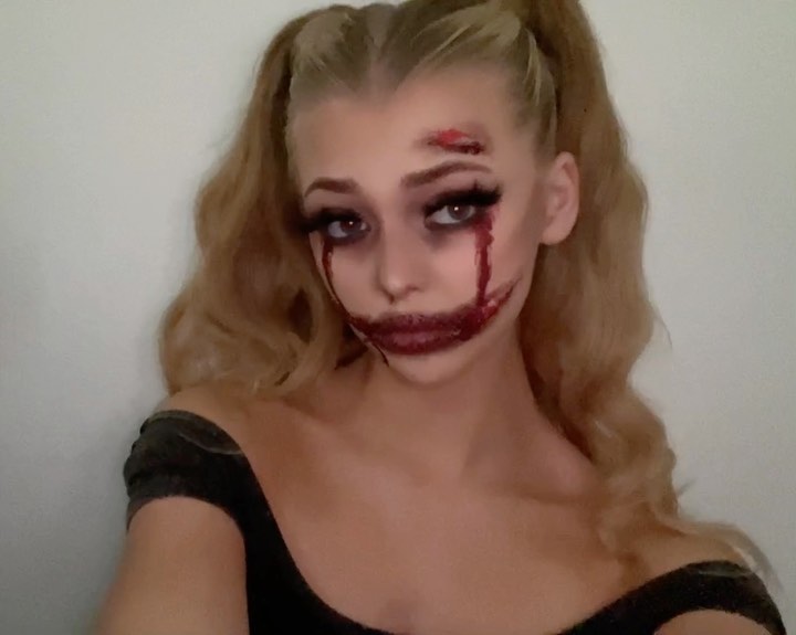 a woman with blonde hair in pigtails is wearing halloween makeup. she has wounds on her forehead and around her mouth, and blood dripping from her eyes and the wounds. her makeup is scary and gory. she is wearing a black dress.