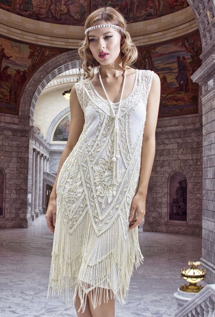 a young woman in a white vintage flapper dress with fringe and beading is standing in a room with stone arches and ornate decor. she is wearing a headband with pearls. she is looking down and to the side, with a wistful expression.