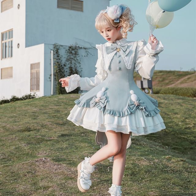 a woman in a blue and white lolita fashion dress is standing on a grassy hill. she is holding blue and white balloons and is looking down at her feet. the background is a white building and a blue sky. the image is taken from an eye level perspective and the lighting is natural.