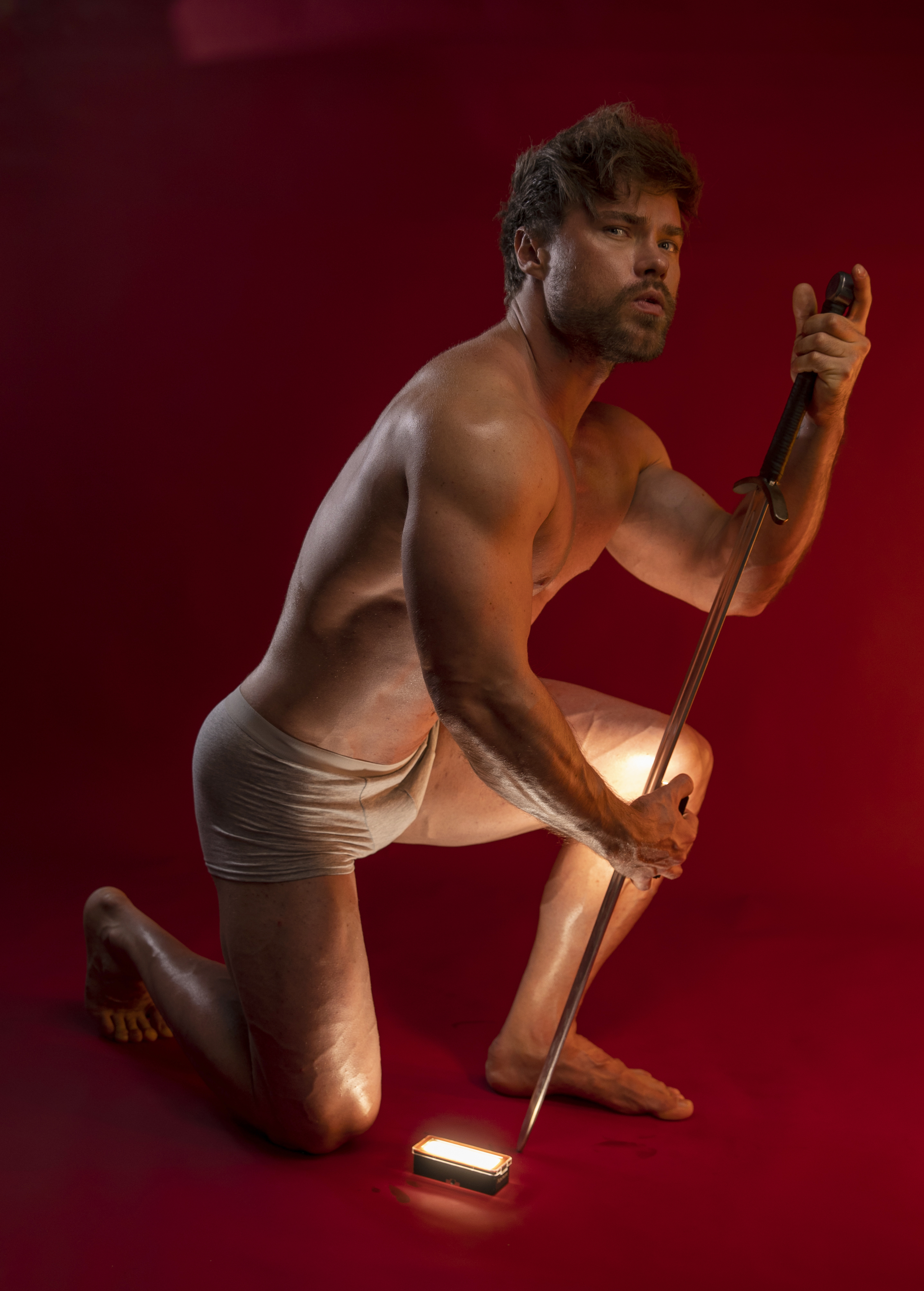a shirtless, muscular male model is kneeling in a studio against a red background. he is wearing lightcolored underwear. he has a beard and is looking at the camera. he is holding a golf club, with the head of the club resting on the ground and emitting a bright light. the model's skin appears to be tanned and oiled.