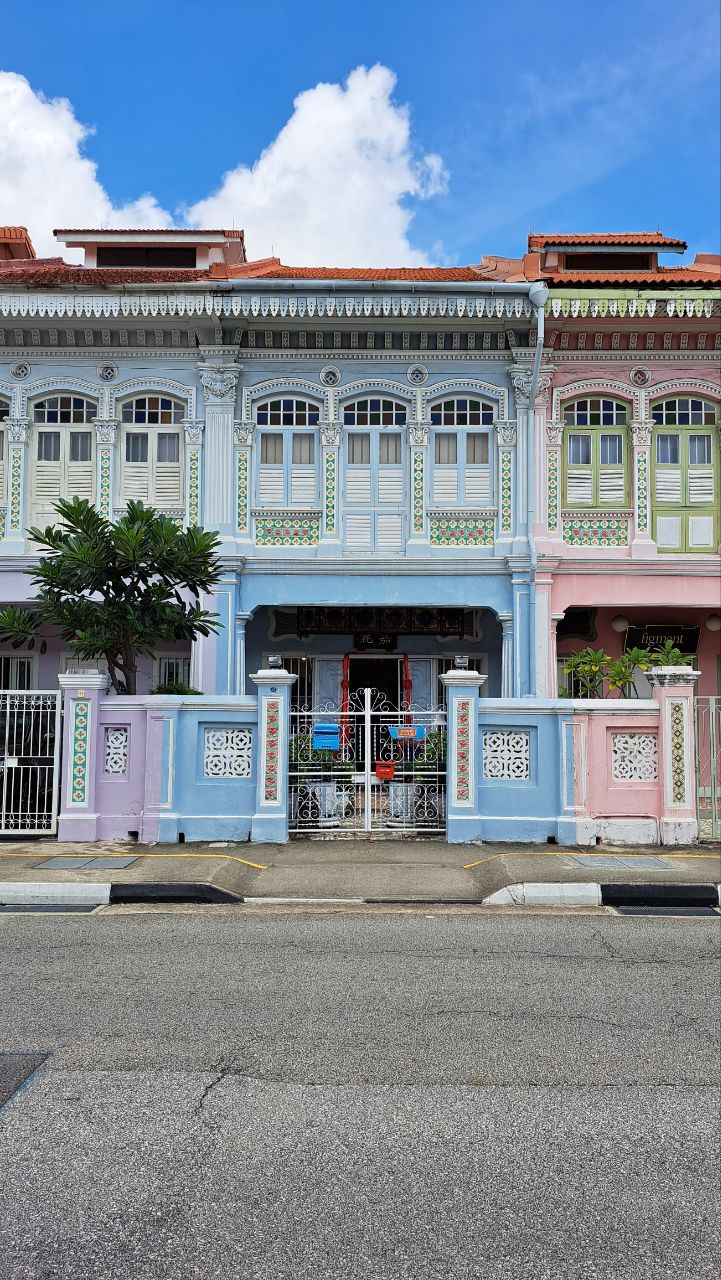 a row of brightly colored shophouses in singapore. the houses are decorated with traditional architectural features, and the street is lined with a sidewalk and a small, black fence. the photo was taken on a sunny day, and the sky is blue with a few white clouds.