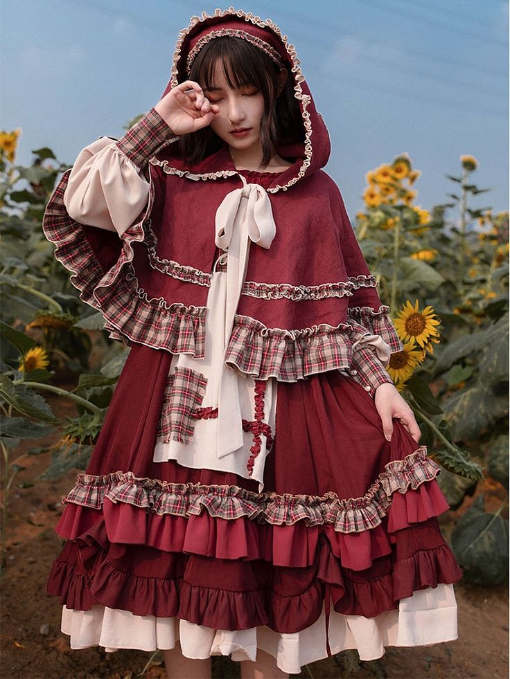 a young woman wearing a red and white lolita fashion dress with a hood stands in a field of sunflowers. the dress has a patchwork design with ruffles and a plaid trim. she is posing with her hand to her head, as if shielding her eyes from the sun. the sunflowers are in bloom and the sky is blue. the overall feel of the image is whimsical and romantic.