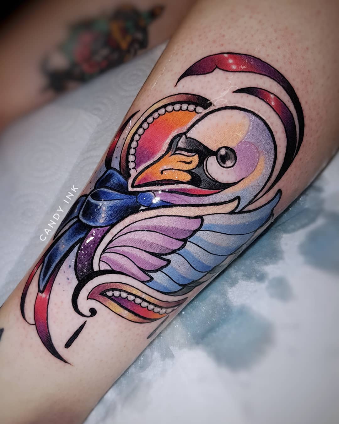 a colorful tattoo of a swan with a blue bow on a person's leg. the tattoo is done in a neotraditional style and features a variety of colors and details. the swan is facing to the right and has its wings folded. the bow is tied around the swan's neck. the tattoo is located on the lower leg, just above the ankle. the tattoo artist's name, "candy ink," is visible in the lower left corner of the image.