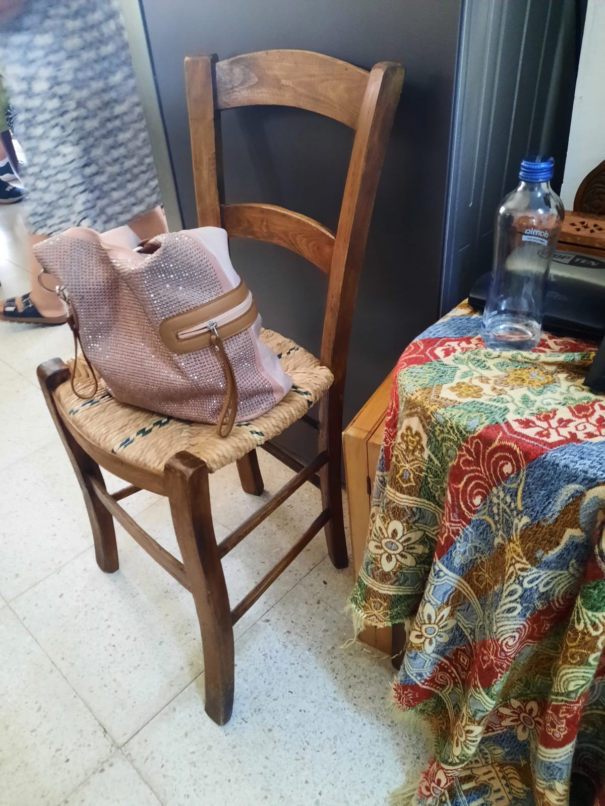a wooden chair with a woven seat has a pink purse sitting on it. a colorful patterned fabric covers a table next to the chair. a water bottle sits on the table. the floor is covered in white tile.