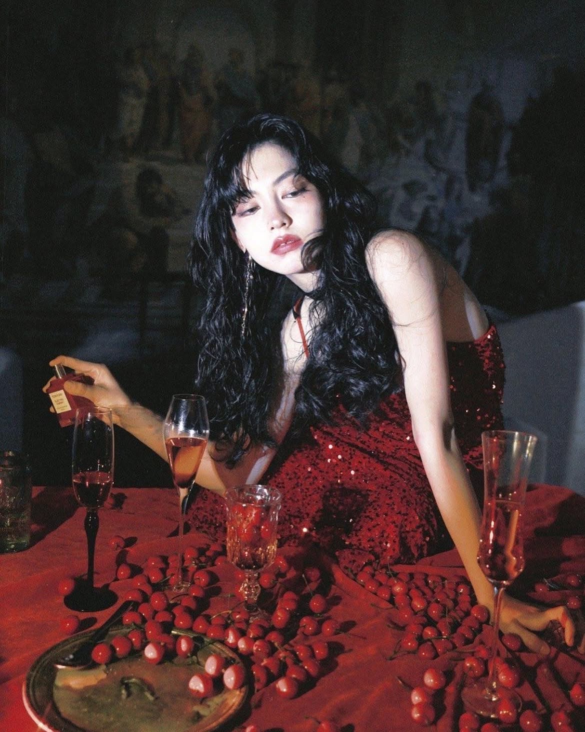 a woman with dark hair and a red dress sits at a table covered with cherries and wine glasses. she is holding a perfume bottle. a painting hangs on the wall behind her.