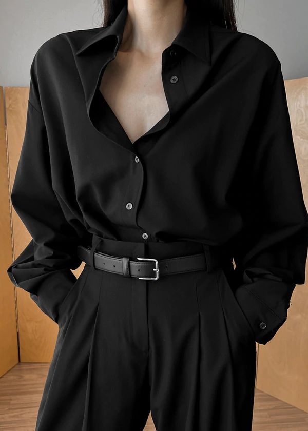 a woman is wearing a black oversized shirt with a black belt and black pants. she is looking at the camera. her hair is pulled back off her face. the background is a muted gray. the image shows a woman in a monochrome, minimal look with a stylish and elegant shirt.