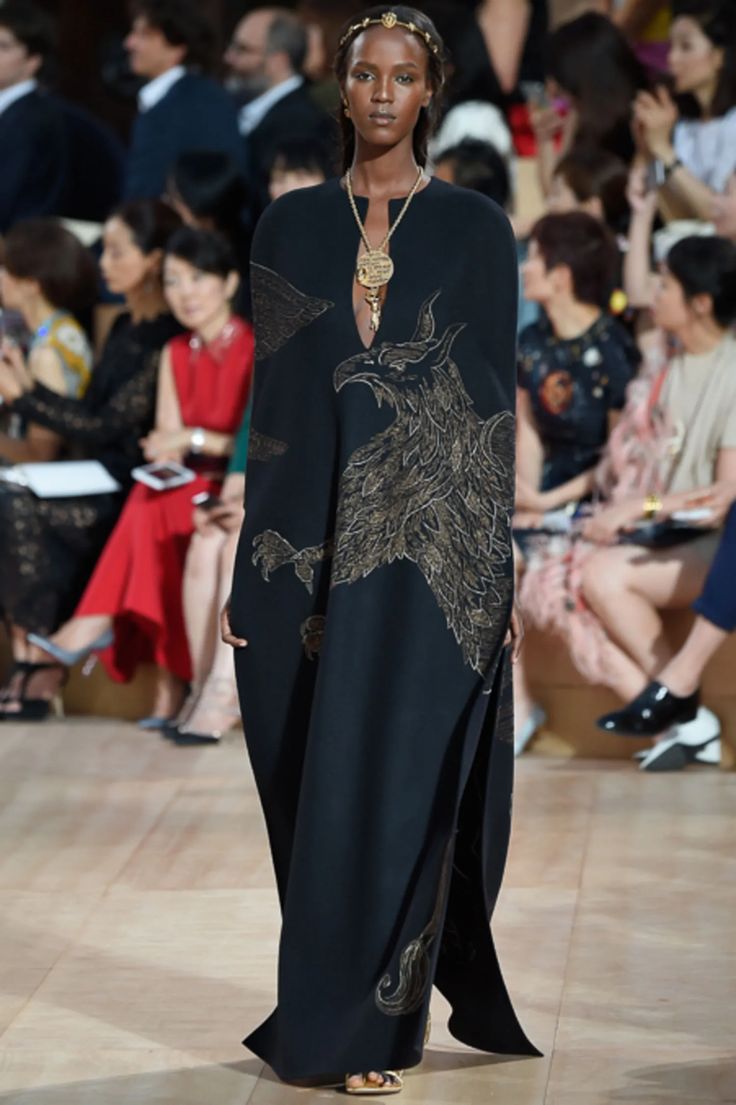 a black floorlength gown with gold phoenix embroidery is showcased on a runway. the model is a tall, slender, darkskinned woman wearing gold jewelry, including a headpiece. the setting suggests a highfashion show, with a blurred background of seated audience members. the dress has a capelike overlay and a high slit. the overall aesthetic is dark, elegant, and sophisticated.