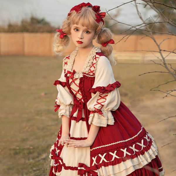 a young blonde girl dressed in a red and cream lolita fashion dress stands in a field with a wooden fence in the background. the dress features intricate lace details, puffed sleeves, and a full skirt. she has pigtails adorned with red ribbons and poses for the camera with a soft smile. the sunlight illuminates the scene, casting warm tones on her outfit and surroundings.