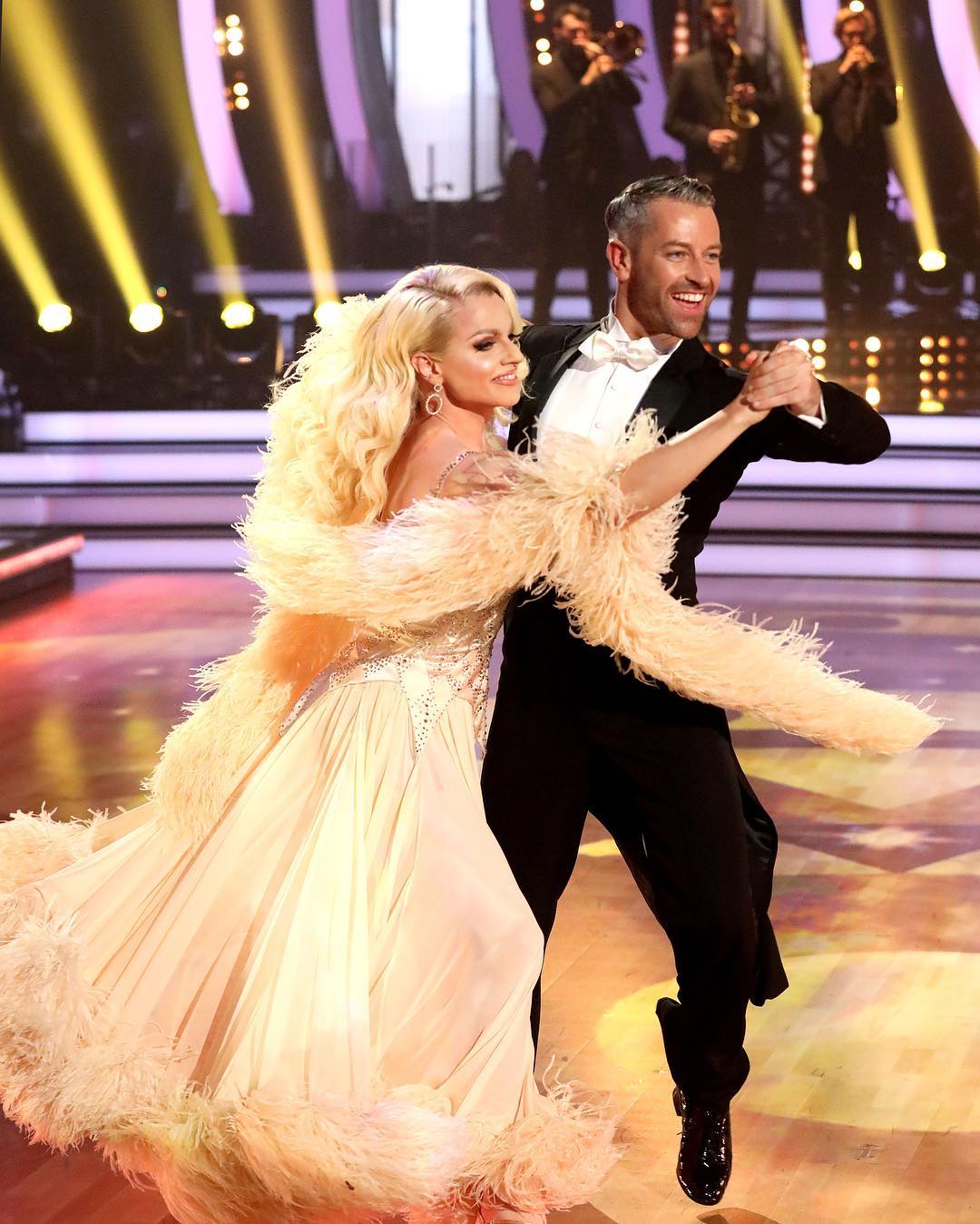 a stunning image captures a ballroom dancing couple in middance.  the woman is wearing a flowing, light beige gown adorned with delicate feathers, while her male partner is dressed in a classic black tuxedo. they are elegantly poised, their movements suggesting grace and precision. the background features a brightly lit stage with a blurred orchestra, creating a sense of a live performance or competition. the overall mood is romantic and sophisticated.