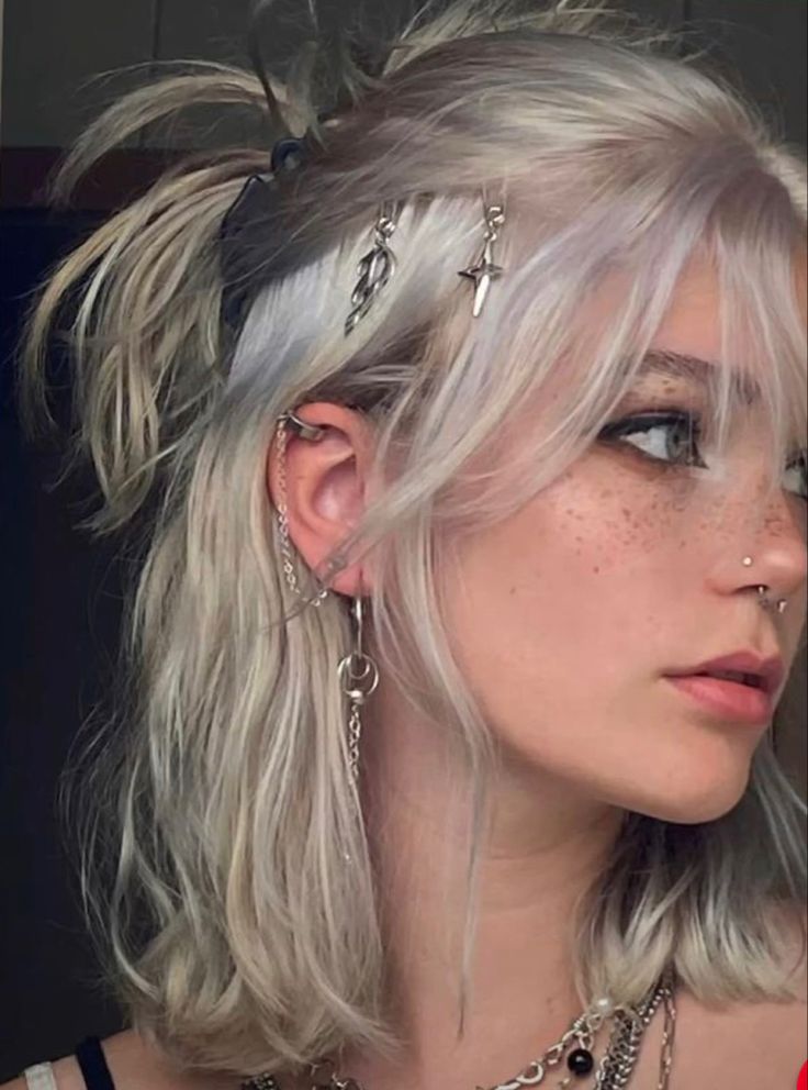 the image showcases a closeup selfie of a young woman with a distinct egirl aesthetic. her short, choppy bob features a blend of silver and light purple hues, styled with a messy, carefree vibe. her makeup highlights a sharp winged eyeliner, enhancing her green eyes, while her complexion remains natural, showcasing freckles. multiple earrings adorn her ears, including a prominent helix piercing with hanging chains. she sports a silver septum ring and a delicate silver necklace. the overall ambiance is softly lit, highlighting the details of her style and features.