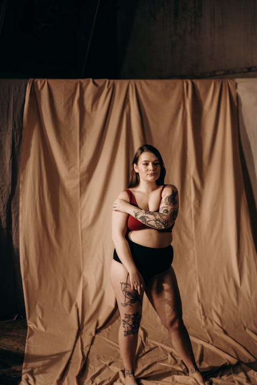 a plussize woman with tattoos stands confidently in front of a beige backdrop, wearing a red bra and black underwear. she is posing with her arms around her body, showcasing her body positivity and selflove. the lighting and setting create a studiolike feel. the image is a celebration of body diversity and acceptance.