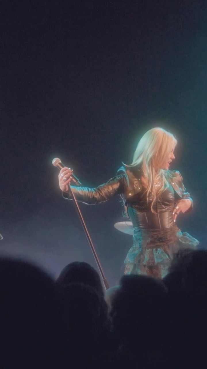 a singer with blonde hair wearing a leather jacket, corset, and ruffled skirt is performing on a stage with a microphone. a crowd is visible in the foreground, and there is stage lighting creating a glowing effect around the singer.
