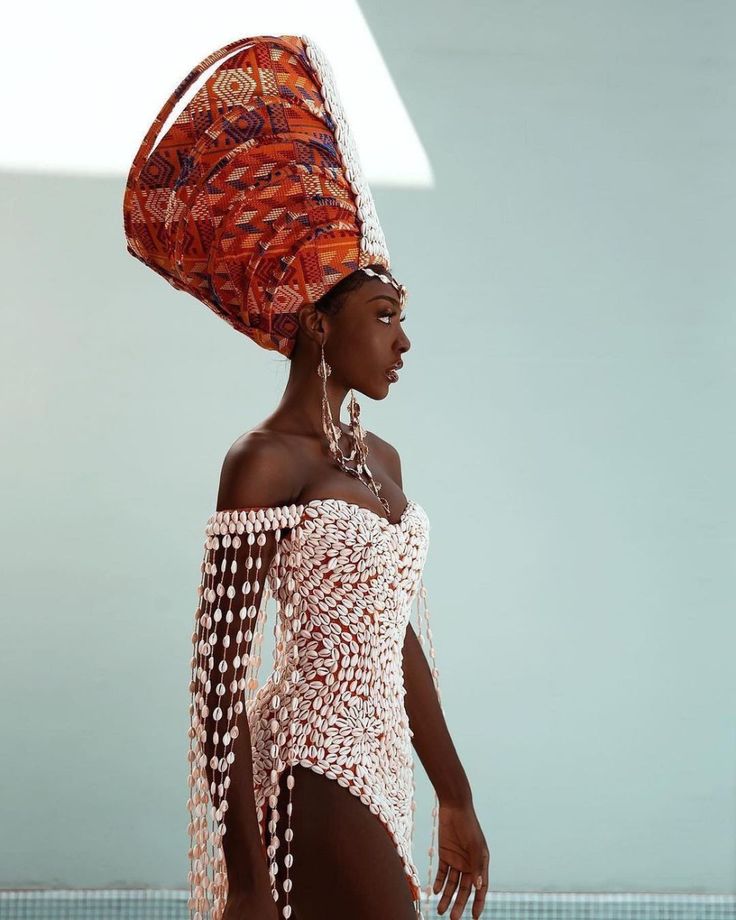 a beautiful darkskinned woman is showcased in a profile view, wearing a striking ensemble. she is adorned with a tall, patterned headdress, a unique offshoulder dress crafted from cowrie shells, and exquisite jewelry including earrings and a necklace. her elegant appearance is set against a light blue background, highlighting her beauty and the intricate details of her cultural attire.