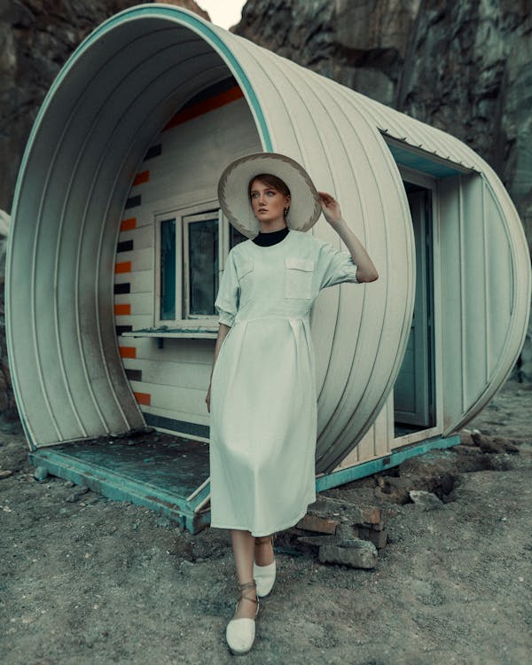 a full shot of a woman wearing a white midi dress and a widebrimmed hat, standing in front of a unique, curved, lightgrey building in a rocky, outdoor setting. the woman is posing slightly turned away from the camera, her expression neutral. she has reddishblonde hair and is wearing white sandals. the building's architecture is unusual, appearing somewhat futuristic or abstract. the overall mood is calm and stylish, with a touch of mystery. the setting suggests a remote or unconventional location.