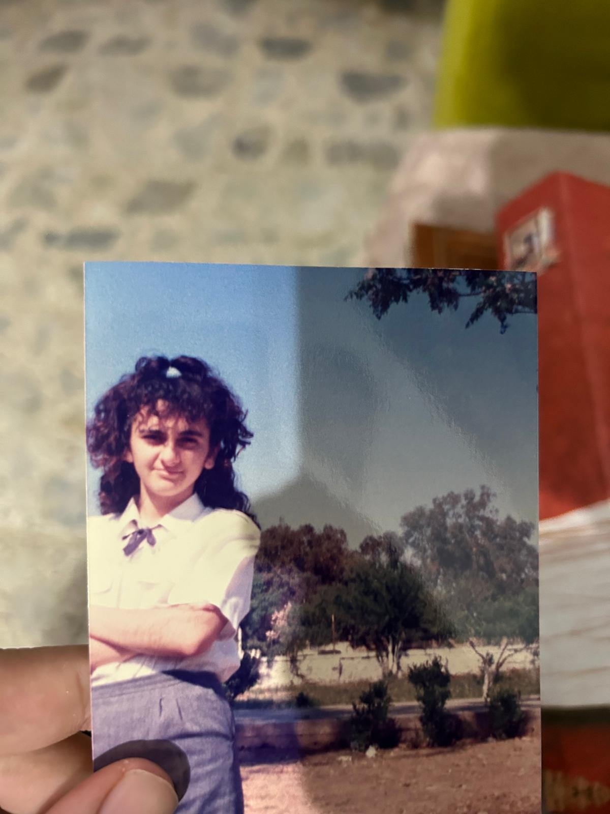 the image shows a closeup of a hand holding a vintage photograph. the photo depicts a young girl, possibly a teenager, with curly brown hair, wearing a lightcolored, collared shirt and a dark gray skirt, typical of a school uniform.  her arms are crossed across her chest. she appears to be standing outdoors in a parklike setting with trees and a sunny sky in the background. the photograph appears to be from the 1980s or 1990s, given its style and quality.  the person holding the photograph obscures a small portion of the image on the left side. a blurry background behind the hand holding the photo shows what appears to be other books or items on a surface.