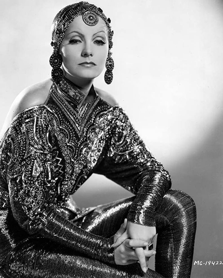 a black and white portrait of greta garbo. she is sitting and looking directly at the camera, with a glamorous expression. she is wearing a beaded gown and jewelry. the image was likely taken in a studio, as evidenced by the plain background.