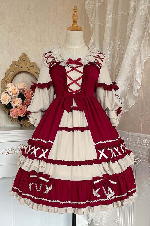 a vintageinspired lolita dress in deep red and cream is displayed on a mannequin. the dress features a high waist, long sleeves with ruffles, and delicate lace trim. a large bow adorns the bodice, which is further embellished with crisscross ribbon detailing. the skirt is full and tiered, with additional lace and ribbon accents. the dress is photographed against a backdrop of white lace curtains and a goldframed mirror, further enhancing its romantic and elegant aesthetic.