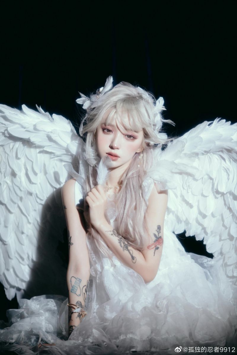 a young woman dressed as an angel with large white wings and a white dress is posing for a portrait. she has silver hair, pale skin, and tattoos on her arms. she is holding a white feather in her hand. the background is dark.