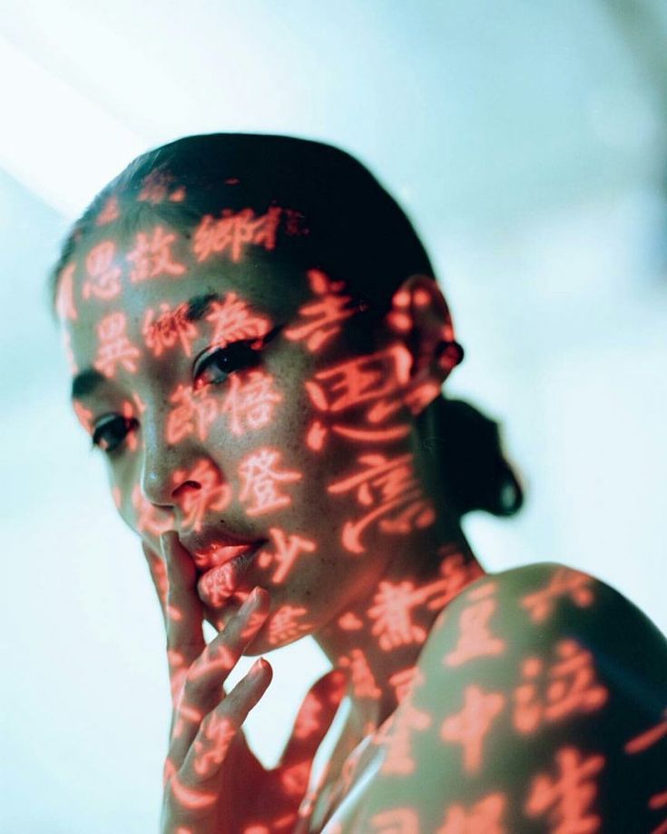 a closeup portrait of a young asian woman with her face covered in projected red calligraphy characters. her hair is pulled back from her face, and she is looking slightly to the left. the background is a soft blue and green. the woman's eyes are closed and her lips are slightly parted. the image has a mysterious and alluring quality.