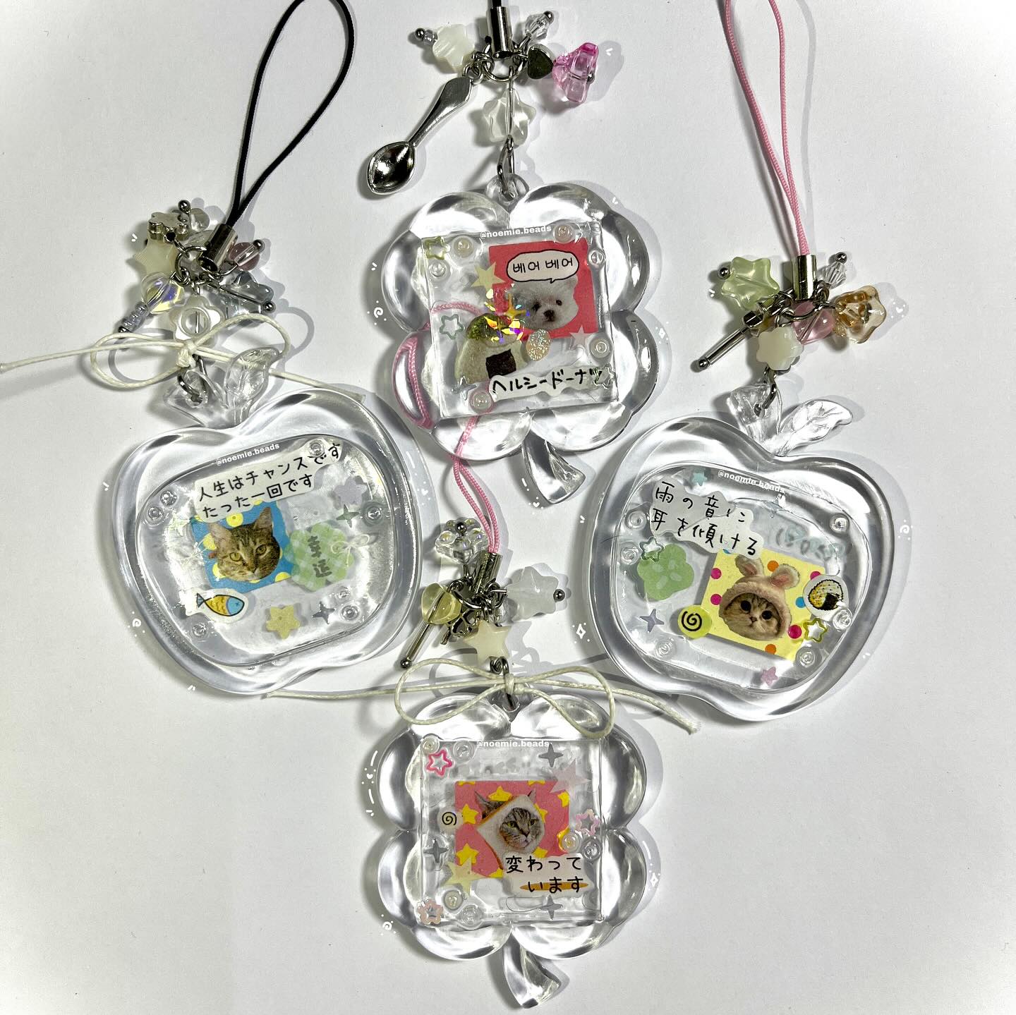 the image shows a collection of clear resin charms or keychains, each containing a small, decorated image inside. some of the charms feature cat images decorated with stickers and text written in both japanese and korean. the charms are shaped like flowers and apples, and they are attached to decorative beads and strings.
