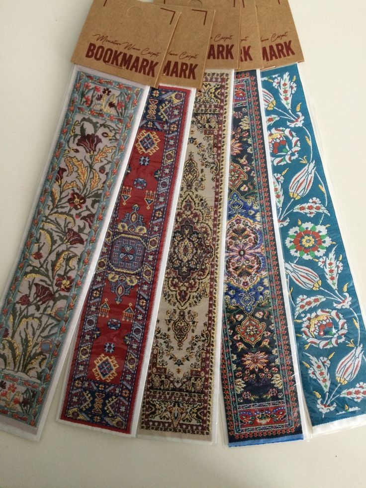 the image shows a collection of five bookmarks arranged in a row. each bookmark has a unique design that mimics the appearance of a miniature oriental rug or carpet. the designs include intricate floral patterns, geometric shapes, and traditional carpet motifs. the bookmarks are made of a thin, flexible material and are housed in clear plastic sleeves. at the top of each bookmark is a brown paper tag with the word "bookmark" printed on it. the designs of the rug patterns range from earthy tones like beige, red, and brown to vibrant colors like blue and turquoise.