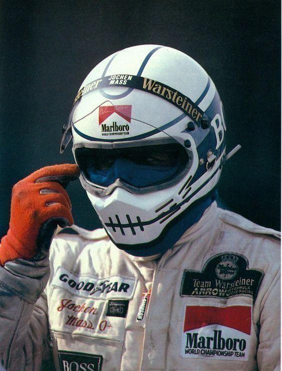 this is a photo of a race car driver in his race suit and helmet. he is wearing a white and blue helmet with black stripes, a white race suit with red, blue and black patches and red gloves. he has a marlboro patch on his suit, and a warstainer patch on his helmet.