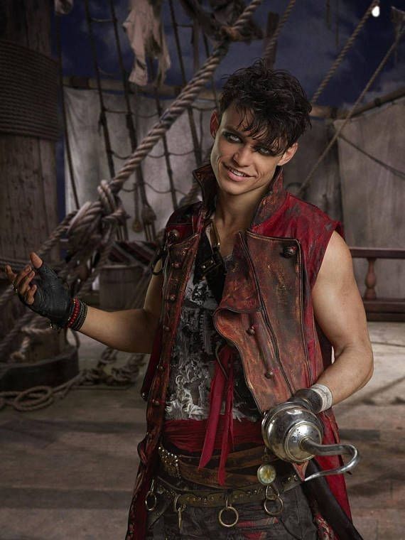 a man dressed as harry hook from the descendants movies smiles at the camera. he is wearing a red vest, a printed tshirt, a red sash around his waist, and several belts. he has a hook on his left hand. the background is a pirate ship set.