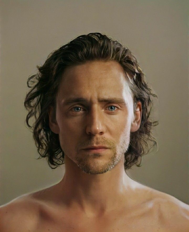 the image is a closeup, highquality portrait of tom hiddleston. he is shirtless, showing his bare chest and shoulders. his hair is long and wavy, brown in color.  he has blue eyes and a serious expression on his face. the background is a plain, neutral beige tone, making the focus entirely on hiddleston.  the lighting and styling suggest a fashion or artistic photography approach.