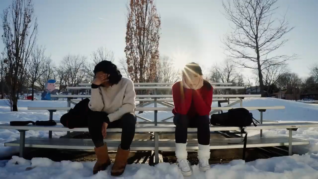two teenage girls sit sadly on snowcovered bleachers in a park.  the sun is behind them creating a backlit effect. the overall tone of the image is somber.