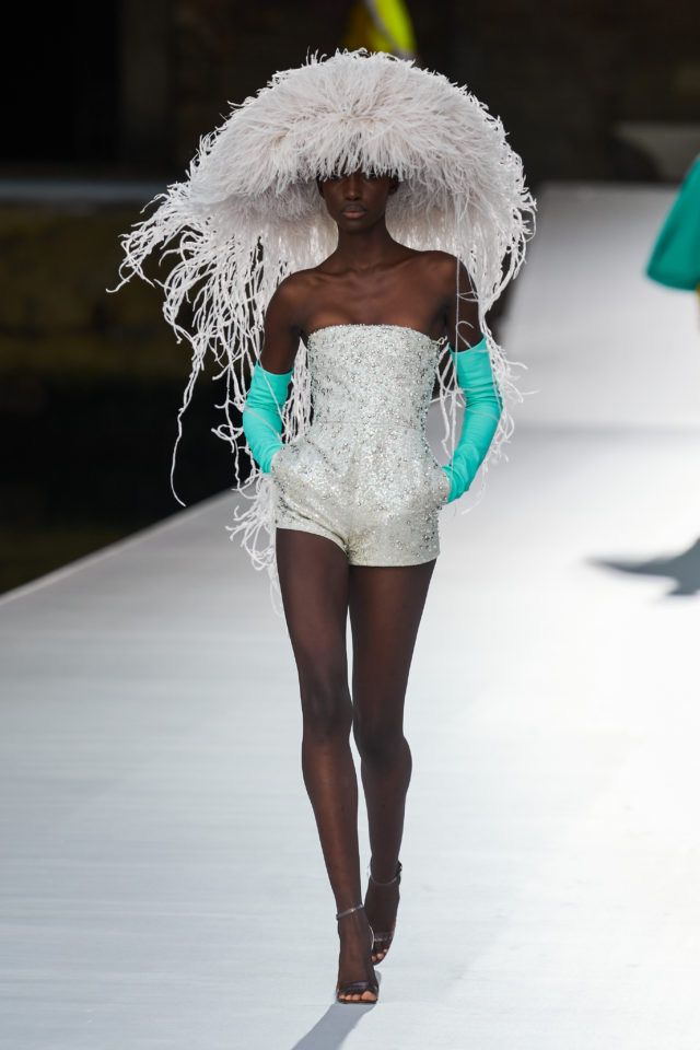 a fashion model walks the runway in a white dress and a large white feathered hat. she is also wearing blue gloves and black heels. the model has dark skin and is posing for the camera.  the runway is white, and the background is blurred.