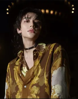 the image is a closeup portrait of a young male kpop idol. he has dark, shoulderlength hair styled with curtain bangs. he's wearing a brown silk shirt with a prominent gold baroquestyle print. the shirt is slightly unbuttoned at the top, revealing a hint of an undershirt. he also wears a thin black choker necklace. the background is dark and moody, with blurred lights suggesting a nighttime setting. the overall aesthetic is stylish and somewhat mysterious. the lighting focuses on the idol's face and upper body, creating a dramatic effect.