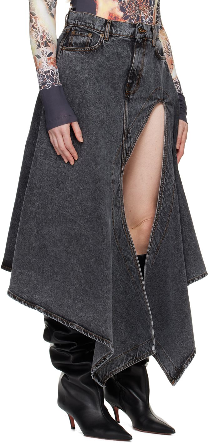 a woman wearing a black denim skirt with a thigh high slit. the skirt is asymmetrical and flows into a point at the back. she is also wearing black thigh high boots.