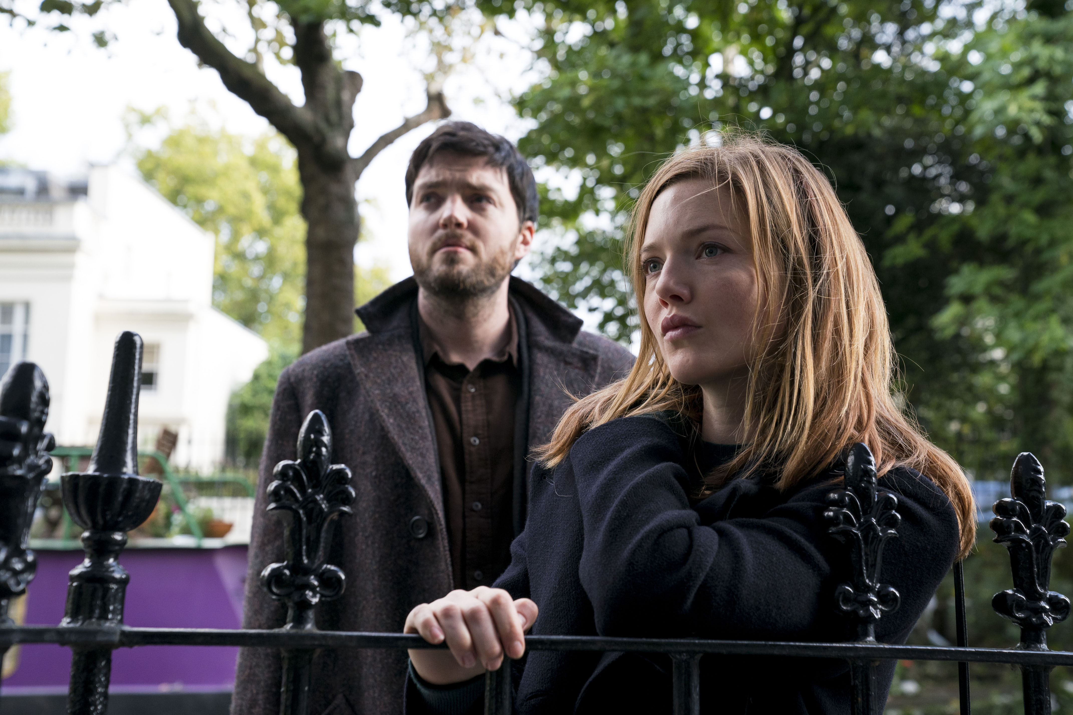 a man and a woman stand together behind a black metal fence in front of a residential building. they appear serious and pensive, as though deep in thought or conversation. the setting is outdoors, in what looks like a residential street in a city. the overall mood is one of mystery or suspense, suggesting a scene from a drama or thriller film or television series.