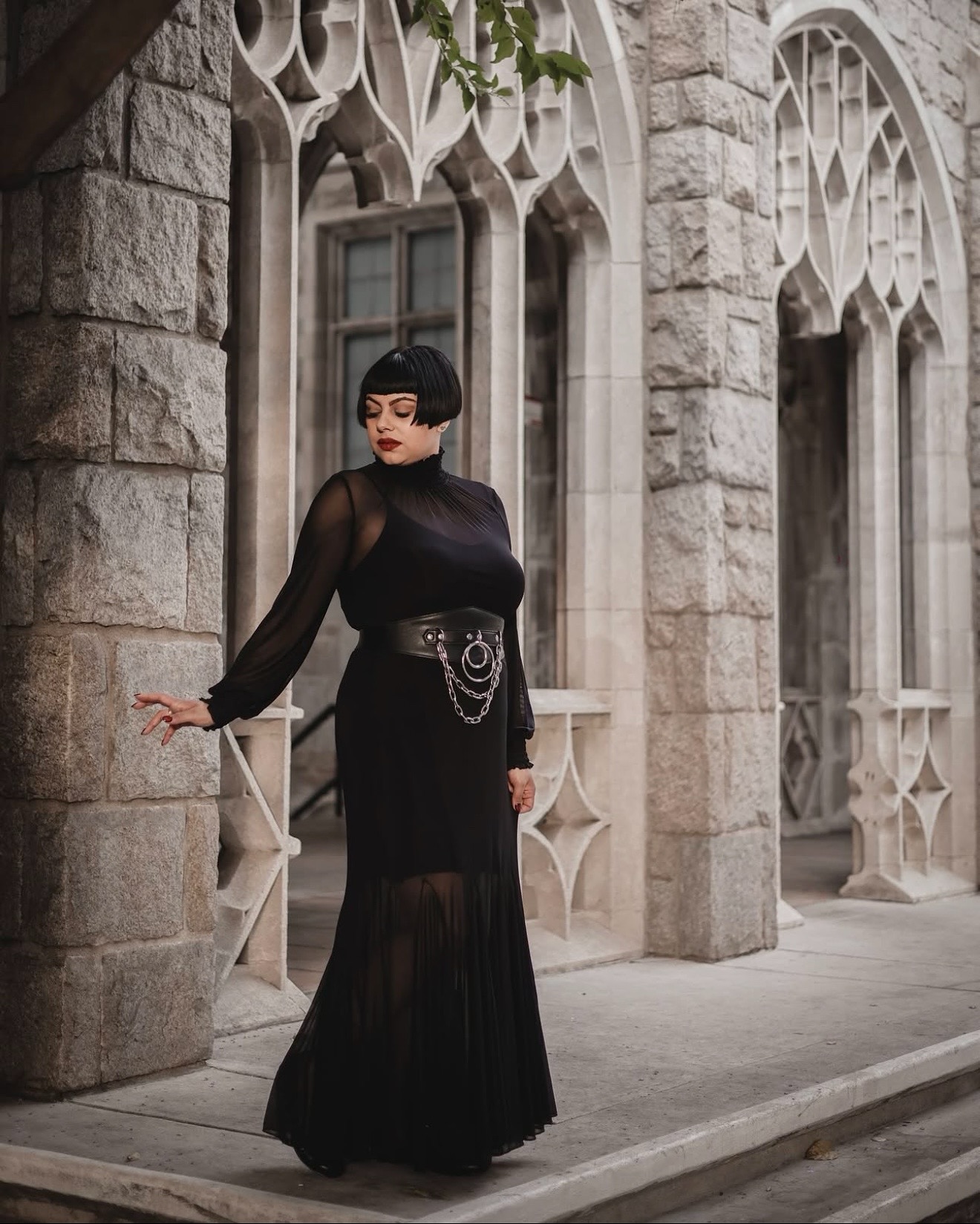 a fulllength portrait of a woman in a black gothicstyle dress. the dress is long, sheer with a pleated skirt, and has a black belt with a chain detail. the woman has short, dark bobbed hair and is posing near a stone building with gothic architecture. the overall mood is dark, mysterious, and elegant.