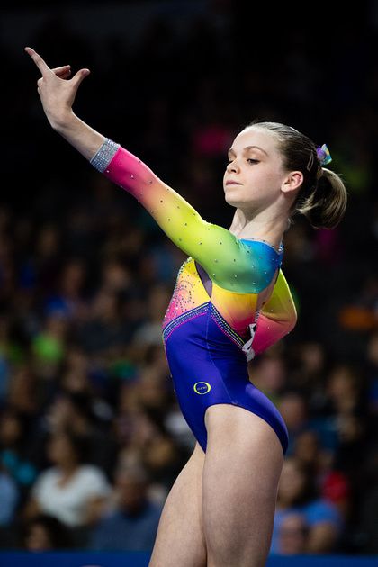 a young female gymnast in a rainbow leotard performs a floor exercise routine with a smile on her face, her arm is extended to the side, and her other hand is on her hip. the gymnast is performing a beautiful and challenging routine, showcasing her flexibility, strength, and grace. the crowd is blurred in the background, but their cheers can be heard.