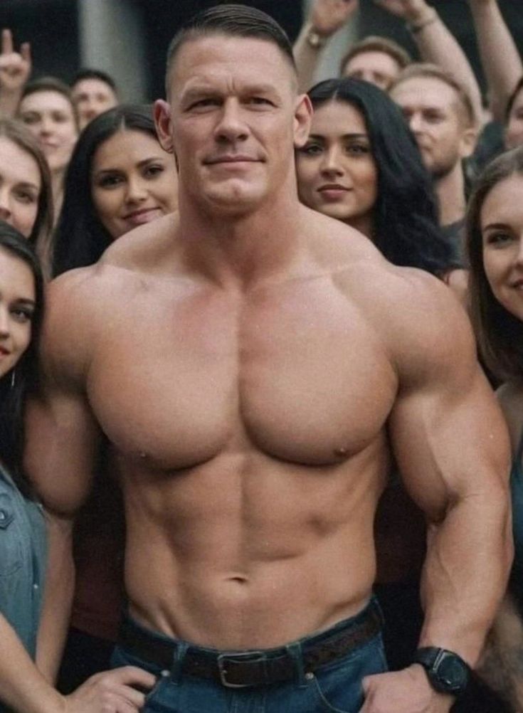 a shirtless john cena is seen smiling while posing for a photo with a crowd of people behind him. john is flexing his right bicep, showing his muscular body with a six pack and sculpted chest.