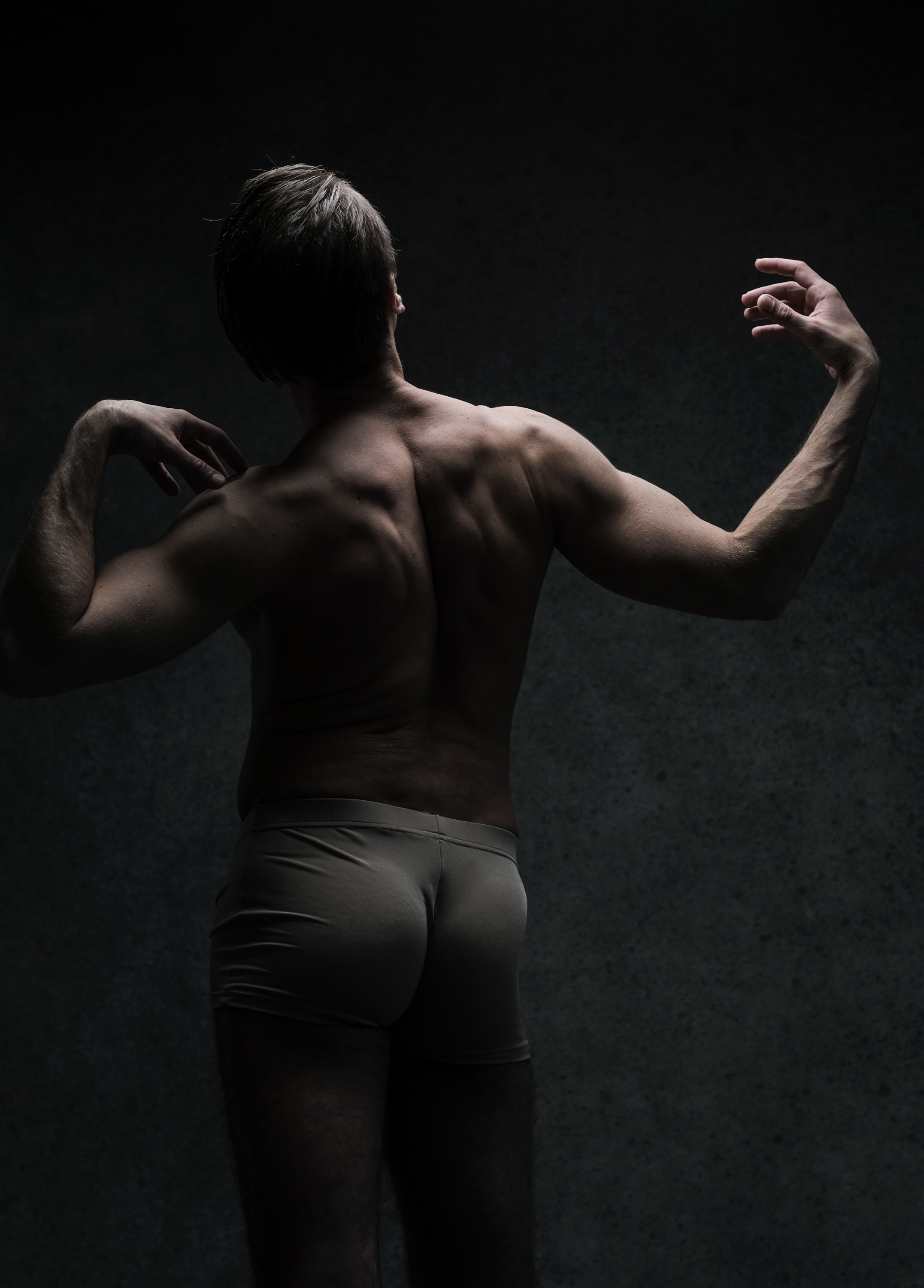 a muscular man in underwear is dramatically spotlit from above, standing with his back to the viewer, twisting his upper body to the right. the background is a dark grey.