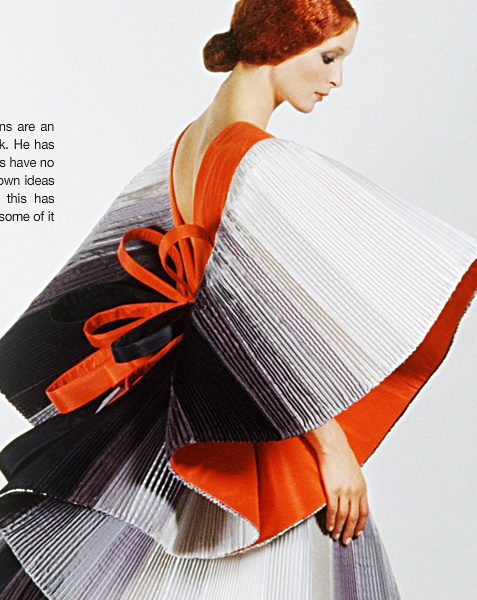 an image showing a woman wearing a unique, vintagestyle dress. the dress is pleated with an ombre effect, transitioning from black at the bottom to white at the top. it features orange accents and an orange bow at the back. the woman has red hair styled in an updo.