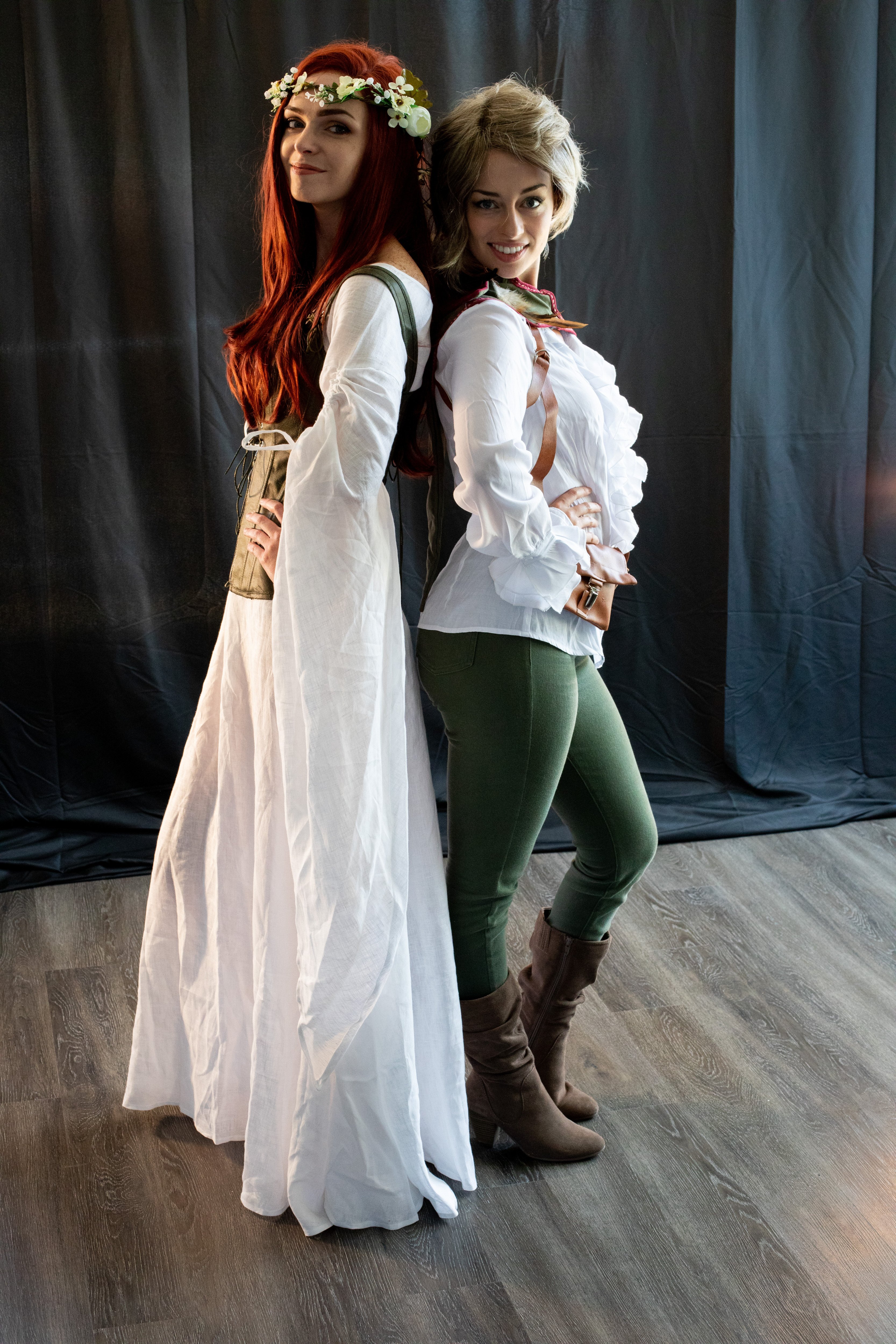 two women stand backtoback in a studio setting against a dark backdrop. the woman on the left embodies an ethereal elf with a white flowing gown, flower crown, and long red hair. the woman on the right portrays a more rugged warrior with a white blouse, green pants, leather accessories, and short blonde hair. their contrasting costumes and appearances highlight the diversity often found in fantasy settings.
