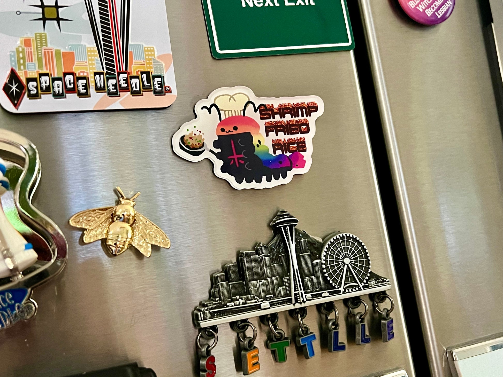 the image shows a stainless steel refrigerator door covered with various magnets and stickers. there's a space needle themed magnet, a cityscape magnet spelling out "seattle", a green "next exit" sign magnet, a round magnet that reads "buzz witch become lesbian", a quirky sticker featuring an anthropomorphic shrimp fried rice dish, and a gold bee magnet.