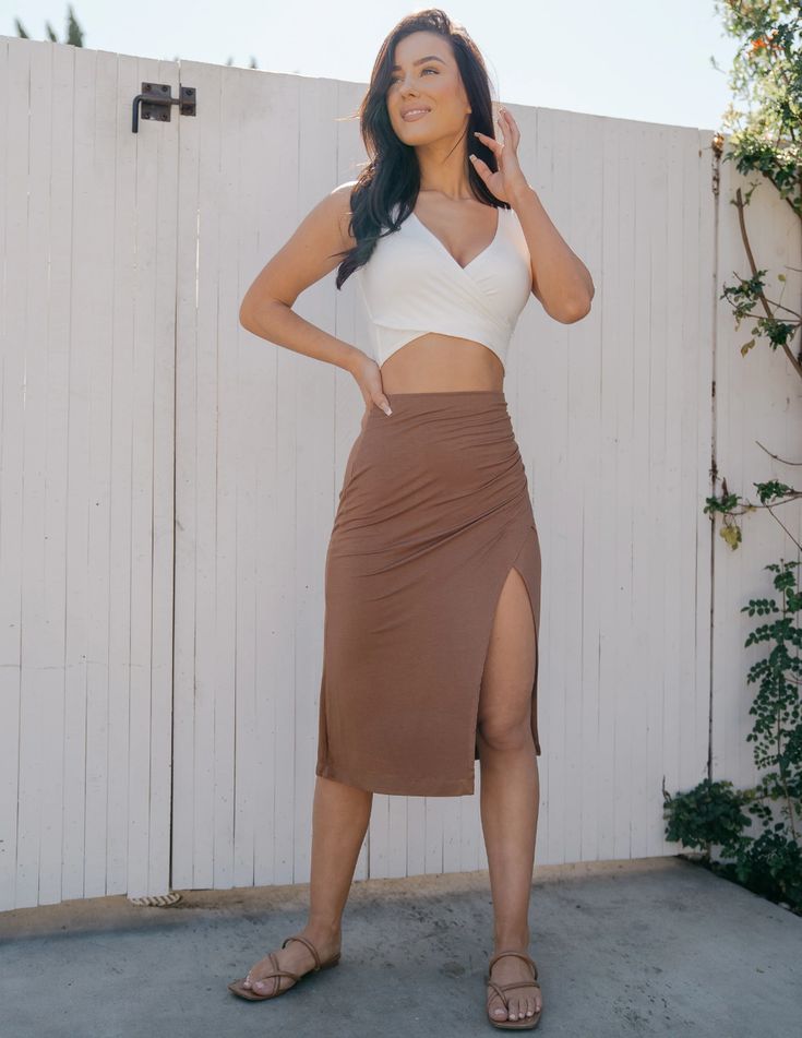a woman is standing in front of a white wooden gate, she's wearing a white crop top and a brown high waisted slit skirt. she's also wearing brown sandals and has her hair down. the image appears to be taken outdoors in natural lighting.