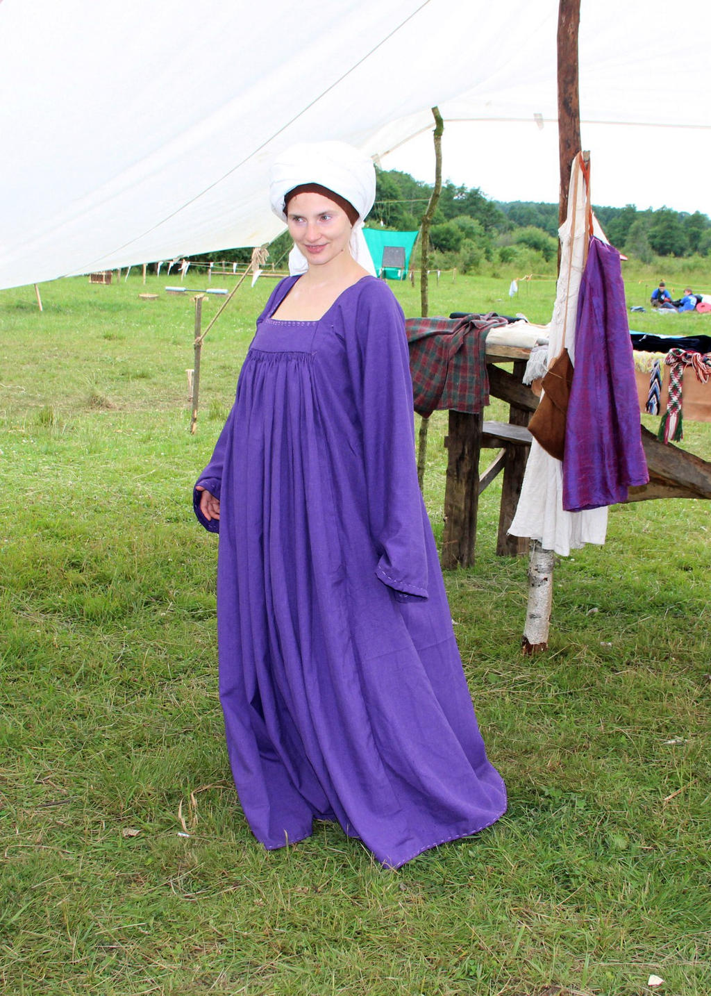 a woman dressed in a long, flowing purple dress and a white head covering stands in a grassy field, possibly at a historical reenactment or medievalthemed event. a camp scene with tents and various items hanging on a wooden structure is visible in the background.