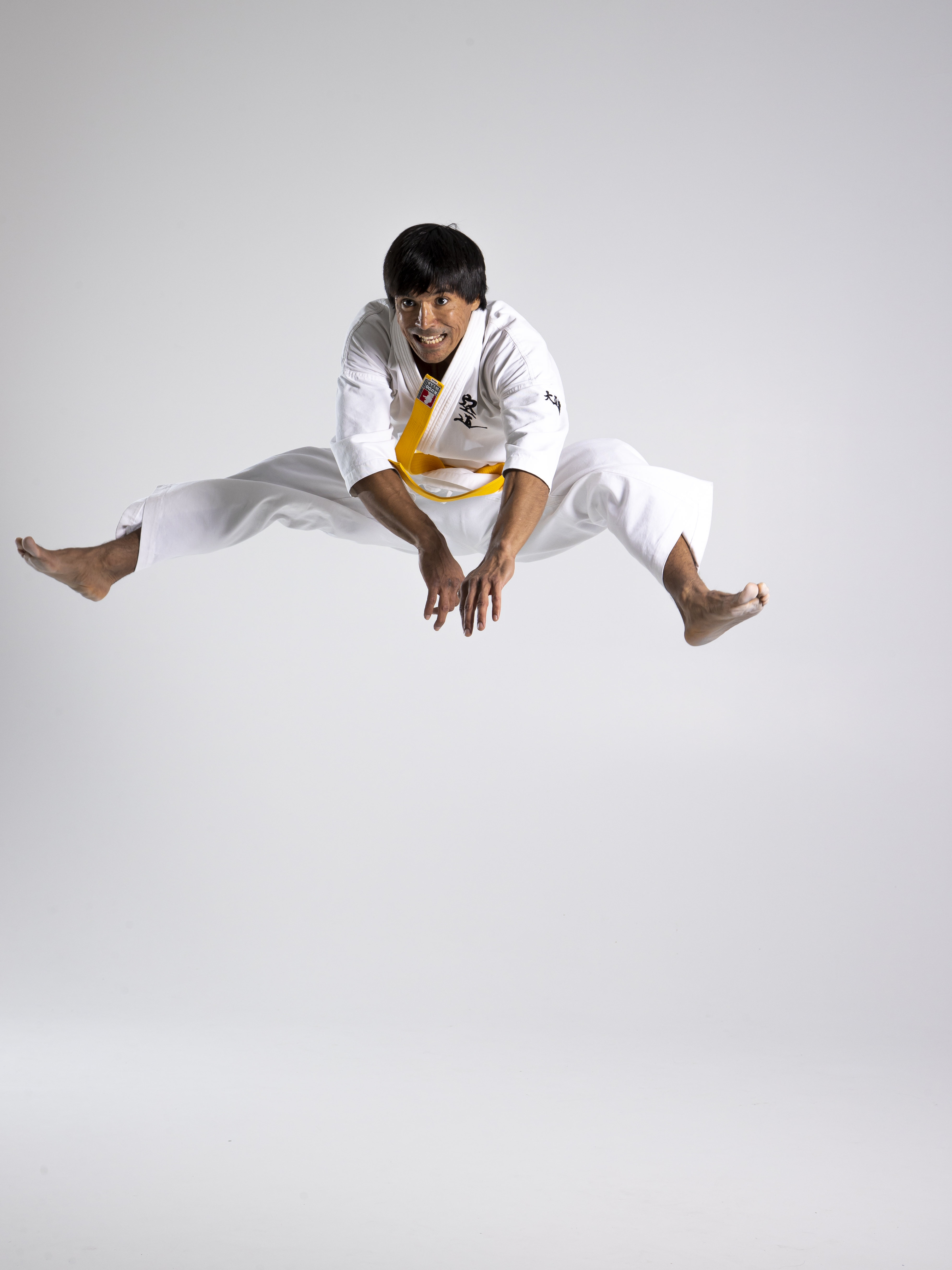 a man in a white karate uniform with a yellow belt is jumping in the air in a wide stance. he is barefoot and the background is a seamless white. he is smiling and his hands are positioned near his knees.